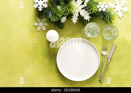 Una vista dall'alto di un tavolo di Natale caratterizzato da un piatto vuoto con candele e posate, abete festoso e decorazioni di alberi bianchi, su un verde luminoso Foto Stock