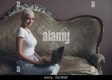 Giovane donna bionda con capelli corti seduti sul divano e sorridente lavorando con il computer portatile. Generazione milleniale business, coaching e blogging concetto Foto Stock