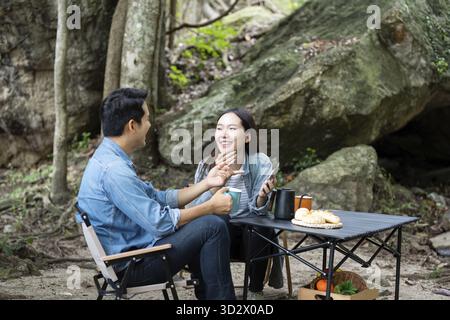 Una coppia ridendo e condividendo momenti con un caffè in campeggio, godendosi la compagnia reciproca in un ambiente naturale Foto Stock