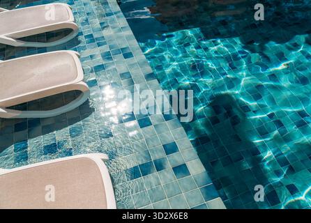 Acqua della piscina azzurra e luminosa accanto a eleganti sedie a sdraio sul bordo piastrellato, che incarnano la vacanza tropicale, il resort di lusso e il relax estivo Foto Stock