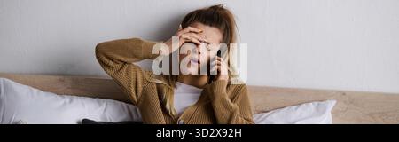 Una giovane donna si rilassa a letto, sentendo il peso del suo esaurimento mentre chatta al telefono, striscione Foto Stock