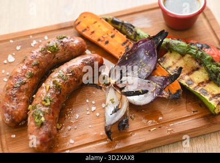 Salsicce grigliate con verdure grigliate su tagliere in legno. Foto Stock
