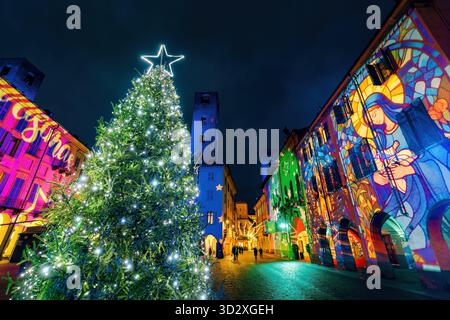 Un albero di Natale si erge illuminato nella piazza, fiancheggiato da edifici adornati da proiezioni luminose, durante il periodo festivo ad Alba, Italia. Foto Stock