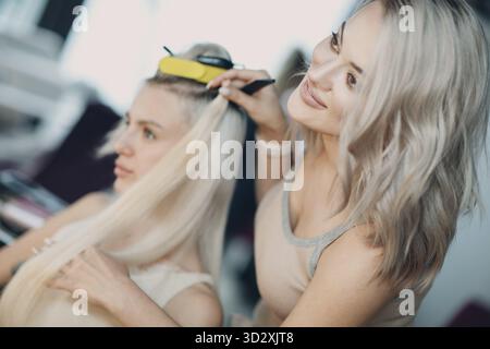 Parrucchiere femmina che fa estensioni di capelli a giovane donna con capelli biondi in salone di bellezza. Prolunga professionale prova controllo Foto Stock