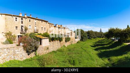 Le storiche case in pietra di Monells, in Catalogna, confinano con una lussureggiante gola verde sotto un cielo blu vibrante in una giornata di sole. Foto Stock