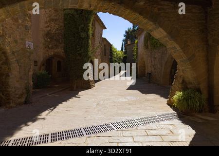 Vista soleggiata di Plaza del oli a Monells, Catalogna, incorniciata da archi in pietra, edifici rustici e vegetazione lungo una tranquilla strada acciottolata. Foto Stock