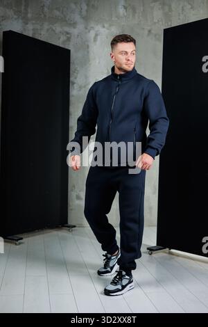 Un uomo alla moda cammina con sicurezza in un elegante outfit atletico, con una giacca con zip e pantaloni coordinati, su uno sfondo minimalista. Lo sguardo è Foto Stock