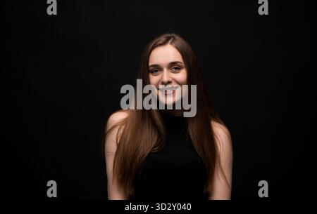 Giovane donna sorridente con lunghi capelli marroni che indossa la parte superiore nera in posa su sfondo scuro Foto Stock