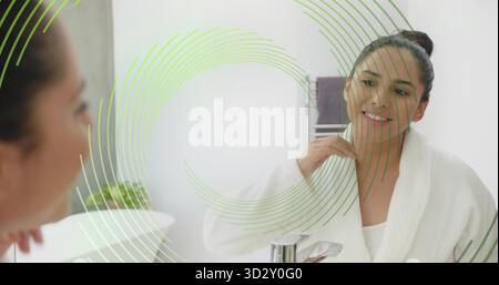 Donna indiana sorridente che indossa un accappatoio bianco che tocca il collo al lavandino del bagno, riflettendo nello specchio Foto Stock