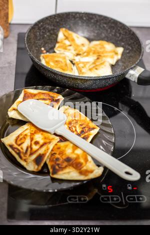 Dolci salati con lavash appena preparati con formaggio fresco su un piatto nero, accanto a una padella su una moderna stufa a induzione. Foto Stock