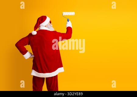 Babbo Natale dipinge una parete gialla con un rullo mentre indossa il suo tradizionale abbigliamento rosso natalizio in una scena vivace Foto Stock