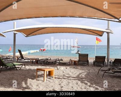 Rilassante spiaggia in Turchia con tettoie ombreggiate, lettini vuoti e un piccolo tavolo di legno su una riva di ciottoli. Acqua blu cristallina, barca lontana e una Foto Stock