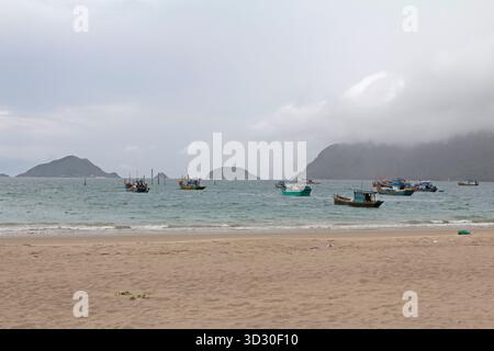 Barche da pesca ancorate al largo in una giornata nuvolosa, viste da una tranquilla spiaggia di sabbia con colline nebbiose dell'isola sullo sfondo. Foto Stock