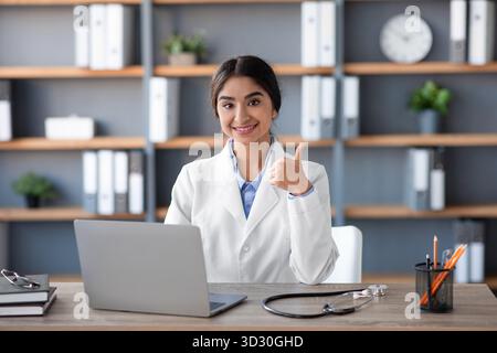 Amichevole indiano donna Dottore in clinica Ufficio dà pollice su mentre si lavora su laptop Foto Stock