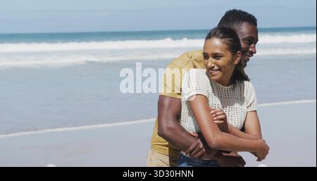 Abbracciare la coppia indossando abiti estivi informali sulla spiaggia sabbiosa ai bordi dell'acqua, con onde oceaniche Foto Stock