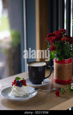 Un'accogliente caffetteria presenta una fetta di torta condita con frutti di bosco, una tazza nera di caffè e un bouquet di fiori freschi, il tutto goduto da una finestra su un su Foto Stock