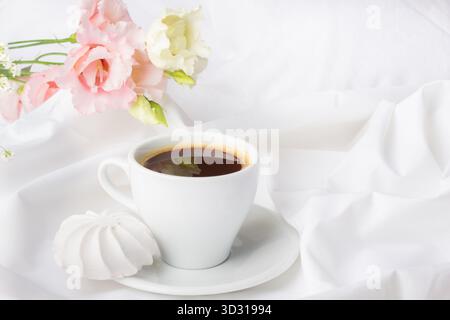 Una delicata tazza di caffè scuro poggia su un piattino bianco. Accanto ad esso, fiori rosa tenui e un biscotto di meringa aggiungono un tocco di eleganza alla serena scena. Foto Stock