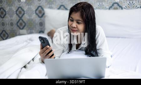 Donna asiatica sdraiata a letto con smartphone e laptop, rilassante a casa, comunicazione online e concetto di stile di vita digitale. Foto Stock