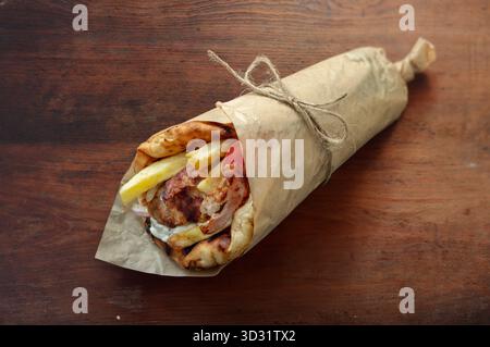 Primo piano di un tradizionale gyros greco con carne alla griglia, patatine fritte e verdure nel pane pita, appoggiato su un rustico tavolo di legno sotto caldo naturale Foto Stock