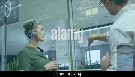 Due colleghi in abiti da lavoro che puntano, guardano le carte e le sovrapposizioni sul divisorio in vetro dell'ufficio Foto Stock