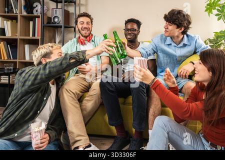 Un gruppo felice e multiculturale di giovani amici (incluso un uomo nero) fa un brindisi con bottiglie di birra mentre si siede su un divano a una festa a casa. Foto Stock