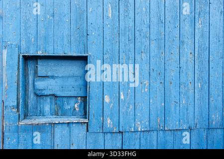 Parete di legno blu intemprata con una piccola finestra quadrata chiusa con tavole rustiche di legno disomogenee St Abbs scottish Borders scozia Foto Stock