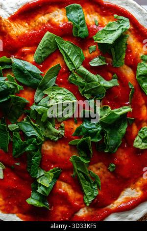 Il processo di preparazione della pizza. Salsa di pomodoro su impasto e foglie di spinaci. Primo piano, vista dall'alto. Sfondo trama Pizza cruda. Foto di alta qualità Foto Stock