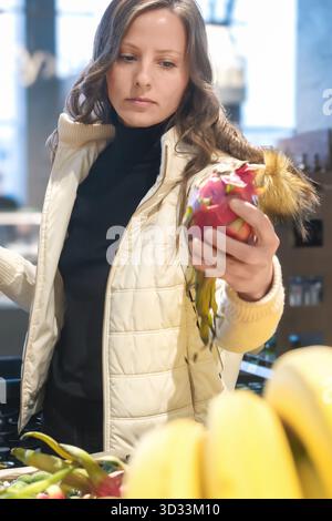 Una giovane donna sceglie la frutta in un negozio, tenendo in mano un frutto Pitahaya rosso. Enfatizza una sana alimentazione mentre acquisti con un cestino Foto Stock