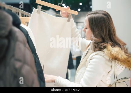 Una donna con una giacca bianca seleziona accuratamente gli abiti dagli scaffali di una boutique elegante, alla ricerca di articoli invernali durante la vendita stagionale in autunno Foto Stock