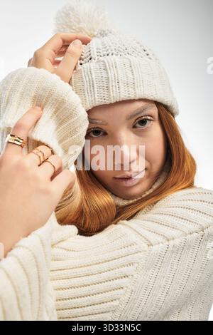 Una giovane donna con i capelli rossi si posa su uno sfondo bianco, indossando un cappello e un maglione in maglia. Foto Stock