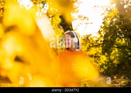 Donna allegra che usa lo smartphone con le cuffie wireless nel soleggiato parco autunnale all'aperto Foto Stock