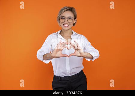Donna romantica che indossa una camicia bianca in stile ufficiale che mostra la forma del cuore con le mani isolate su sfondo arancione Foto Stock
