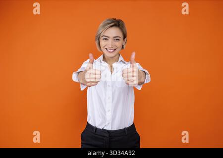 Donna positiva che indossa una camicia bianca in stile ufficiale che mostra gesti e sorride alla fotocamera isolata su sfondo arancione Foto Stock