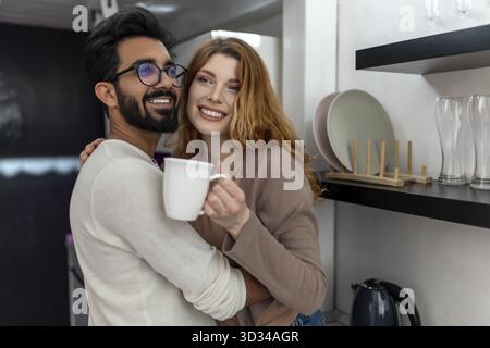 Donna gioiosa che beve un caffè con un uomo e si abbraccia mentre si siede sul bancone della cucina Foto Stock