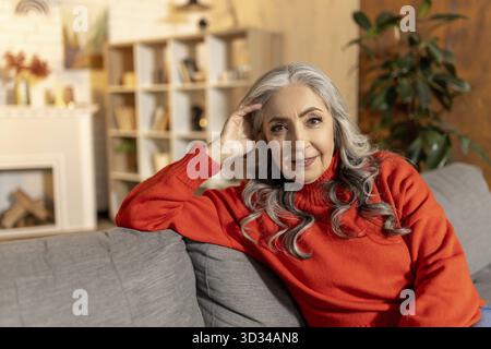 A casa. Bella donna anziana dai capelli lunghi seduta sul divano a casa Foto Stock