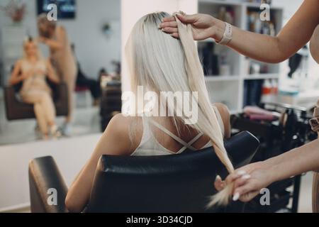 Parrucchiera femmina che fa le estensioni dei capelli alla giovane donna con i capelli biondi nel salone di bellezza. Salone professionale di estensione dei capelli Foto Stock