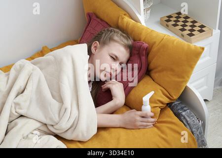 Il bambino sembra malato e triste. Ragazza che usa spray medico volare a letto a casa, mal di gola Foto Stock