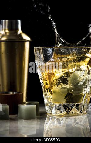 Whisky alcol spruzzare in vetro sulla barra, shaker in background, scarsa illuminazione Foto Stock