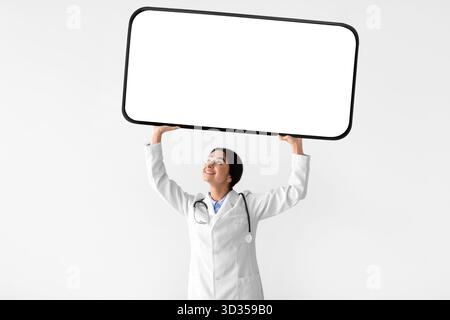 Felice donna indiana Dottore in cappotto bianco tiene grande telefono con schermo vuoto sopra la sua testa Foto Stock