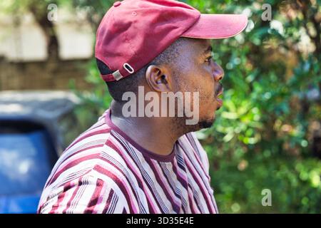 un giovane uomo africano in sovrappeso che cammina nel cortile, indossa una t-shirt a righe e un berretto Foto Stock