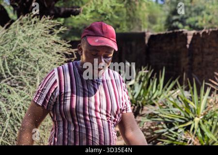 un giovane lavoratore africano in sovrappeso che cammina nel cortile, indossando una t-shirt a righe e un berretto Foto Stock