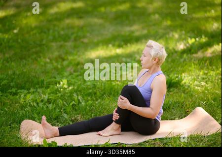 Una donna di mezza età medita mentre siede su un tappetino per il fitness in erba all'aperto. La donna esegue strette all'anca, praticando movimenti consapevoli durante l'esterno Foto Stock