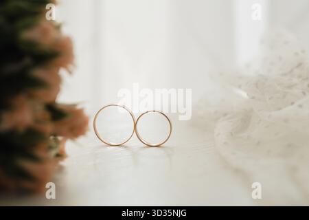 Due semplici fedi nuziali dorate poggiano su una superficie leggera, accanto a fiori delicati e tessuti fluenti, a simboleggiare l'amore e l'impegno in un ambiente interno sereno Foto Stock