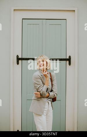 Una donna dai capelli biondi corti sorride gioiosamente fuori da una porta azzurra. È vestita con un elegante abito grigio con pantaloni bianchi, che trasuda conf Foto Stock