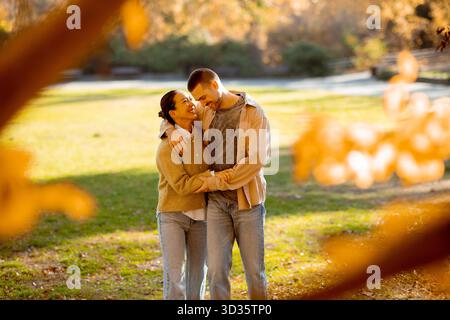 Una gioiosa donna coreana e un uomo caucasico condividono un momento di amore in un bellissimo parco, circondato da vibranti colori autunnali e dalla luce soffusa del sole. Foto Stock
