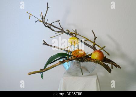 Una disposizione floreale unica con ramoscelli, fiori gialli luminosi e una pesca, presentata su un piedistallo minimalista su uno sfondo bianco. Il de Foto Stock