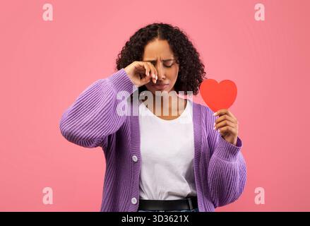 La giovane donna in Purple Cardigan tiene il cuore rosso mentre piange per problemi di relazione Foto Stock