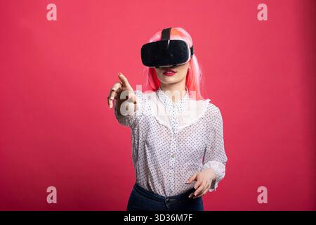 Giovane donna con parrucca rosa che indossa un visore VR, punta il dito verso la fotocamera durante l'interazione con un ambiente 3D coinvolgente. La curiosa donna caucasica esplora il mondo digitale futuristico. Foto Stock
