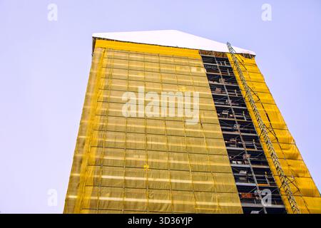 Edificio torreggiante in costruzione si erge alto, avvolto da impalcature gialle che si affacciano sul cielo blu. Il suo tetto bianco di picco contrasta nettamente con Foto Stock