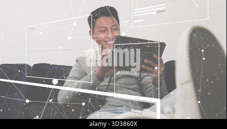 Uomo sorridente in felpa con cappuccio che tiene il tablet appoggiato sul divano scuro di casa, con grafica di rete Foto Stock